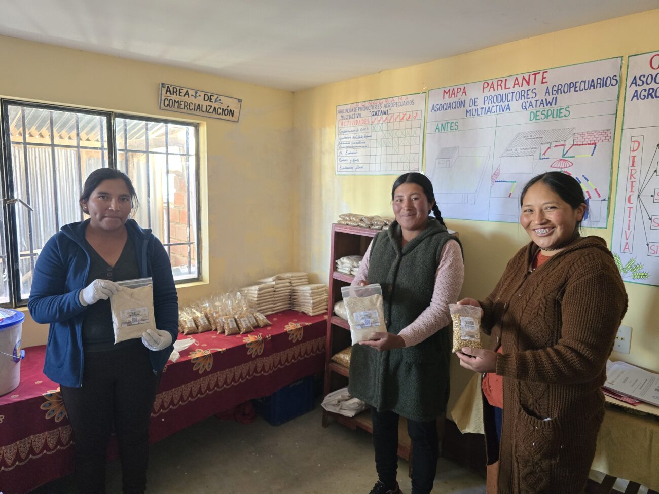 Tres productoras de Q’ATAWI sostienen bolsas transparentes con cereales procesados y granos. Detrás de ellas se observan más productos envasados, estantes y materiales informativos de la organización.