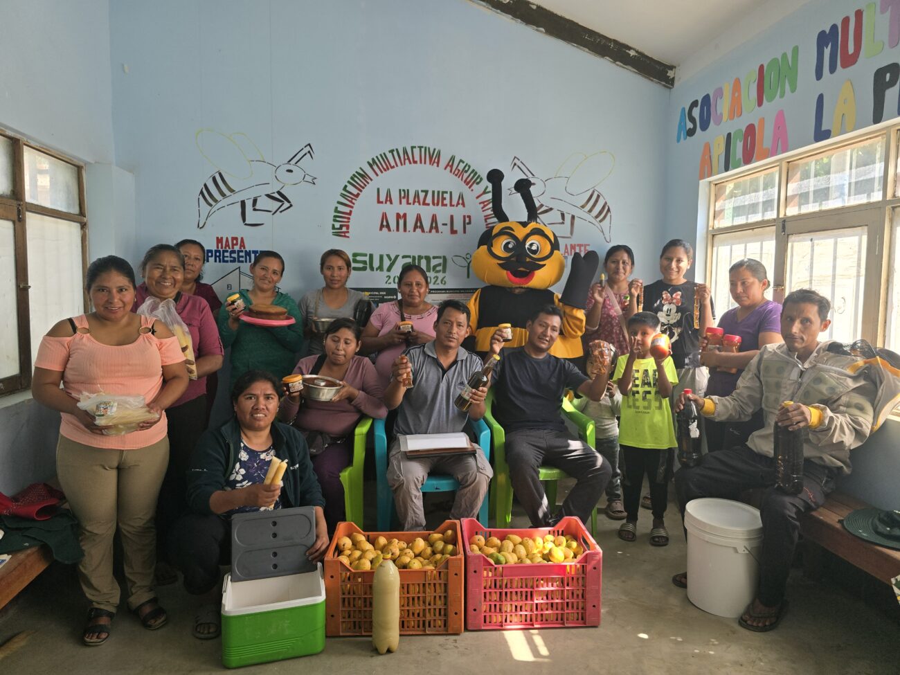 Grupo de mujeres, hombres y un niño posa en un ambiente de AMAA-LP sosteniendo productos elaborados. Al frente hay dos cajas con mangos y, al centro, una persona con un disfraz de abeja.