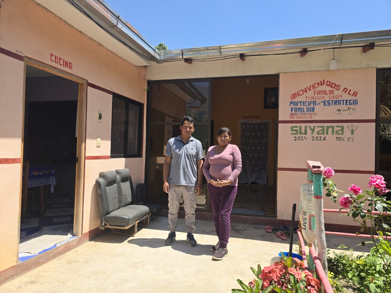  Un hombre y una mujer posan en el patio de una vivienda de color rosado. En la pared se observa un mensaje que identifica a la familia Tangara Quispe como participante de la Estrategia Familiar, junto a las puertas señalizadas de la cocina y el dormitorio.