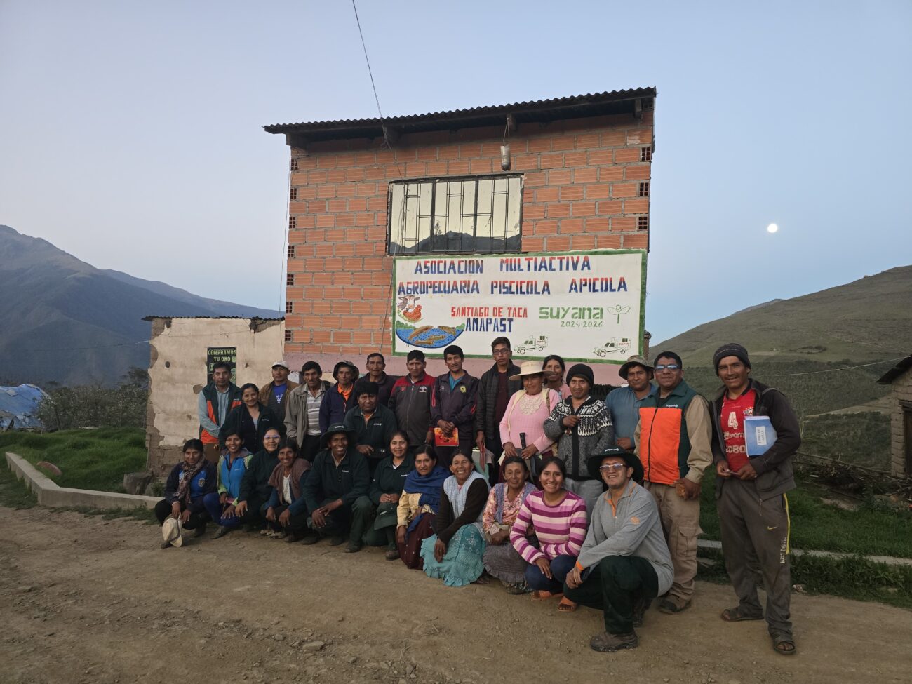 Grupo de mujeres y hombres posa frente a la sede de la Asociación Multiactiva Agropecuaria Piscícola Apícola Santiago de Taca (AMAPAST), en una comunidad rural rodeada de montañas.