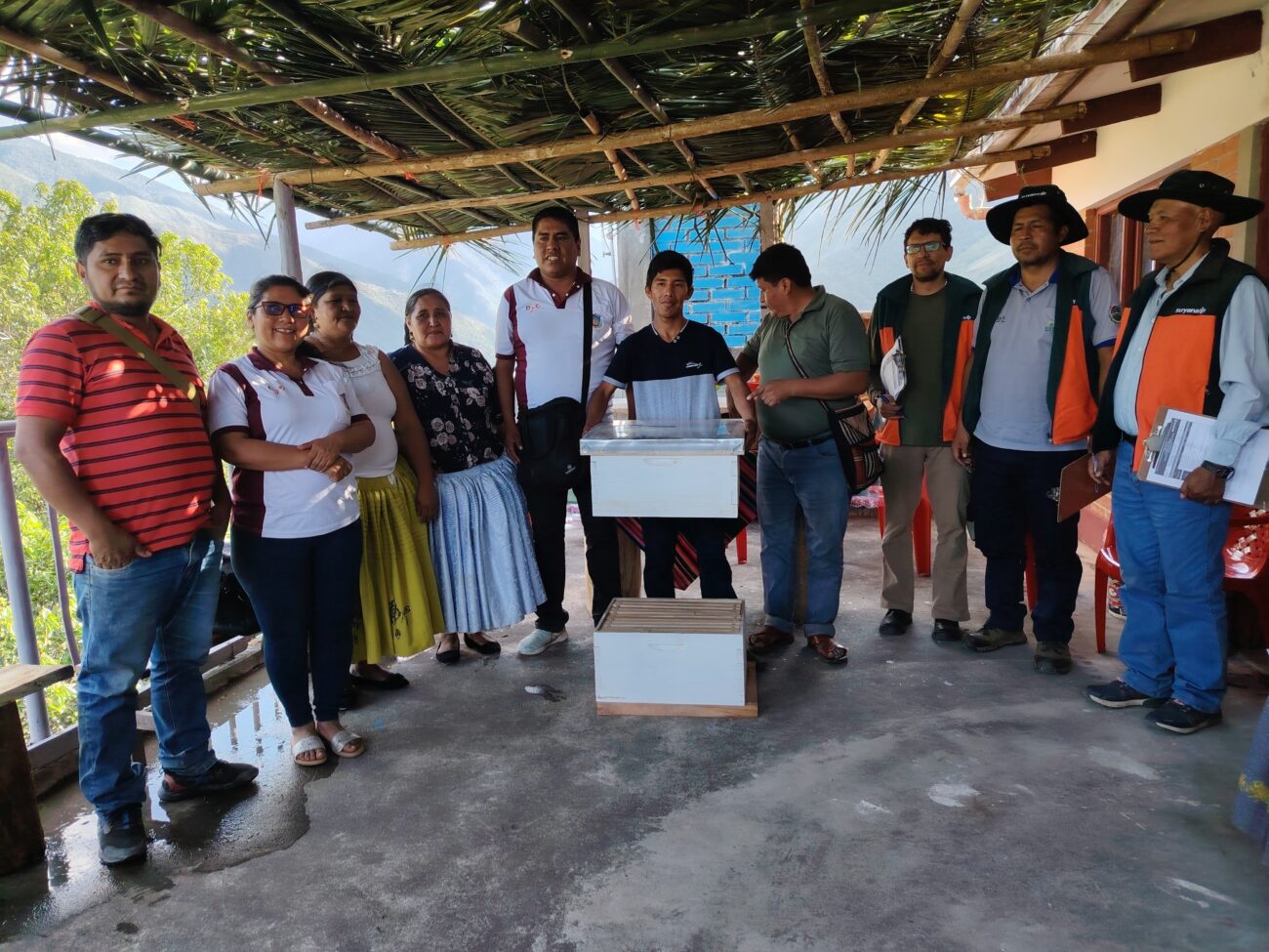 Grupo de mujeres y hombres de la OECA AIAYM, junto al comité evaluador, posa bajo un techo de palma. En el centro se observan dos cajas blancas para colmenas y, al fondo, un paisaje montañoso.